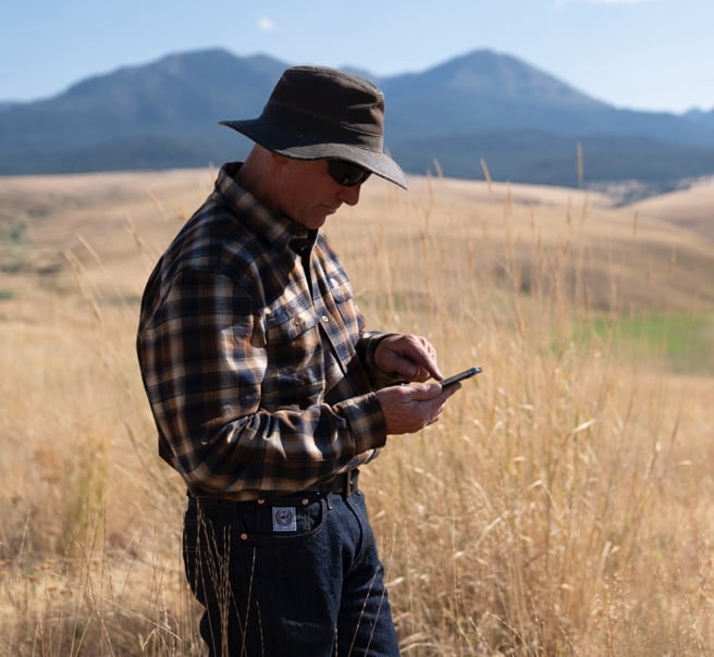 A farmer stands in a pasture and uses a touch screen device.