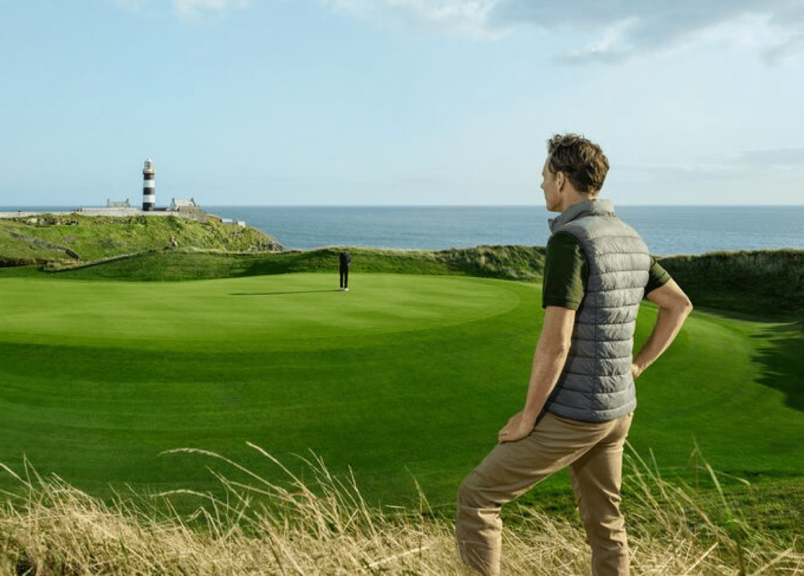 Man overlooking a golf course green and distant lighthouse on the ocean shoreline