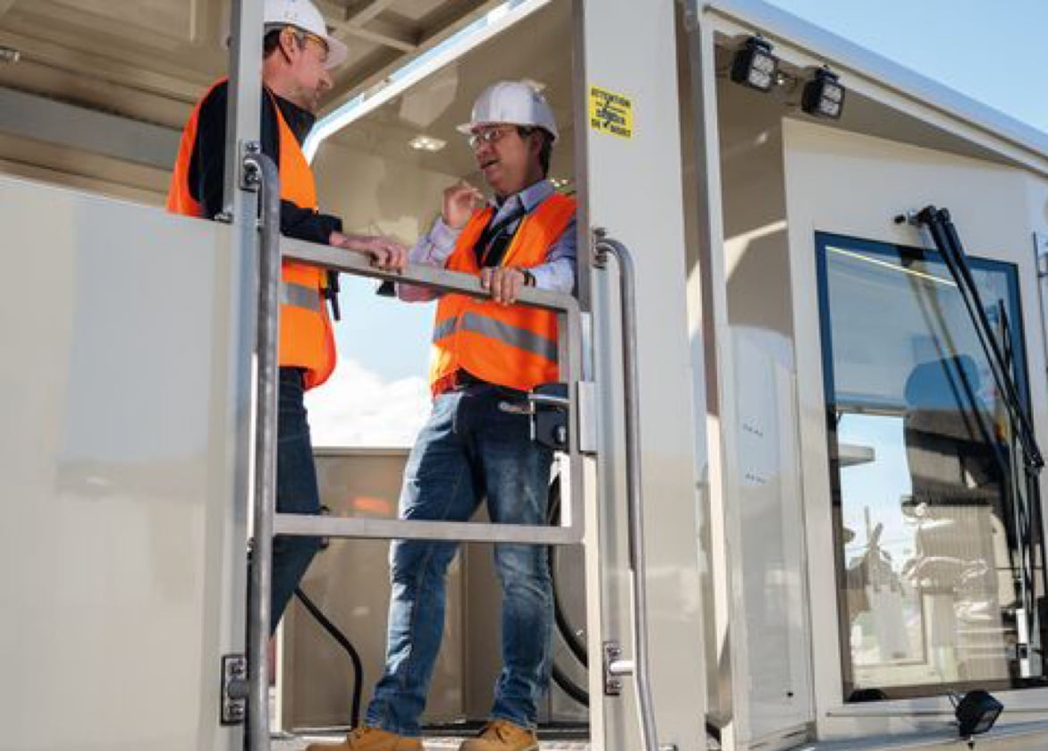 Men wearing high visibility vests and hard hats talk while standing inside vehicle