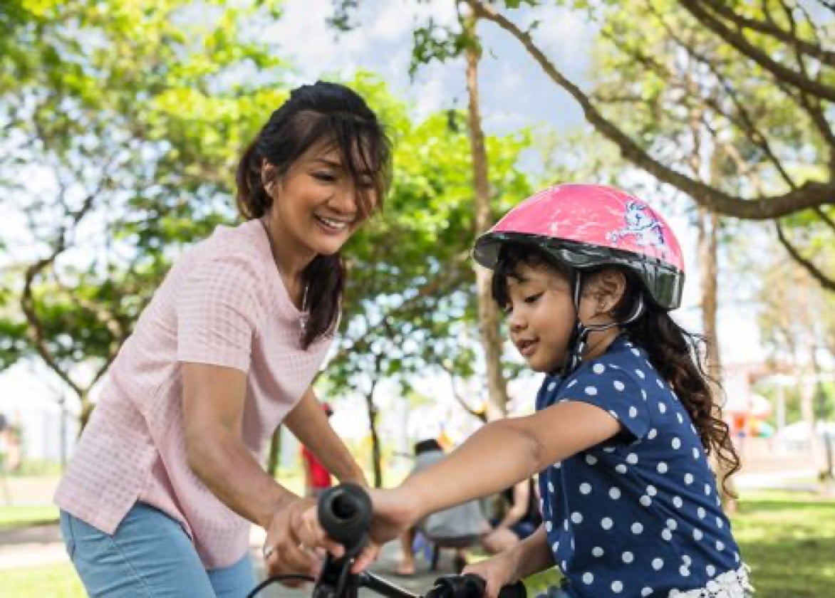 A child wearing a helmet rides a bike while a woman holds the handlebars.