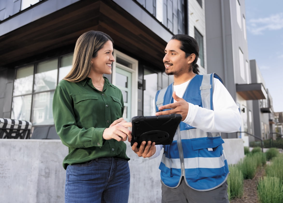A man wearing a work vest and holding a tablet talks to a woman in front of a house.