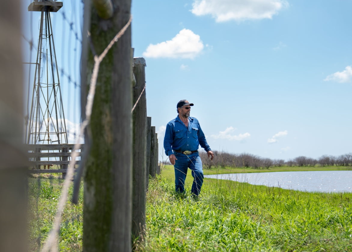 A man wearing a baseball cap walks along a wood and wire fence and looks out over a body of water. 