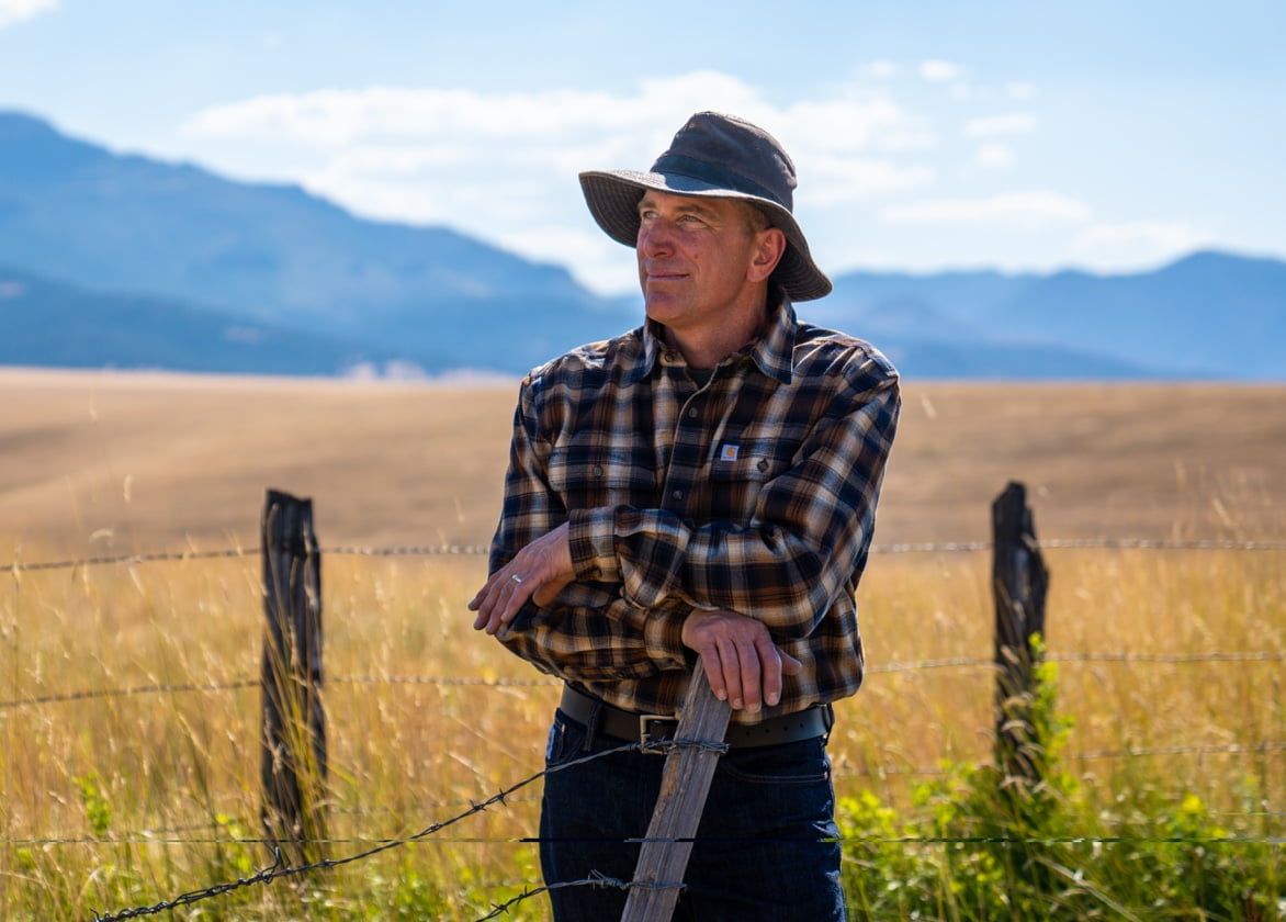 A farmer leans against a wooden fence post and smiles into the distance with a pasture and hills behind him.