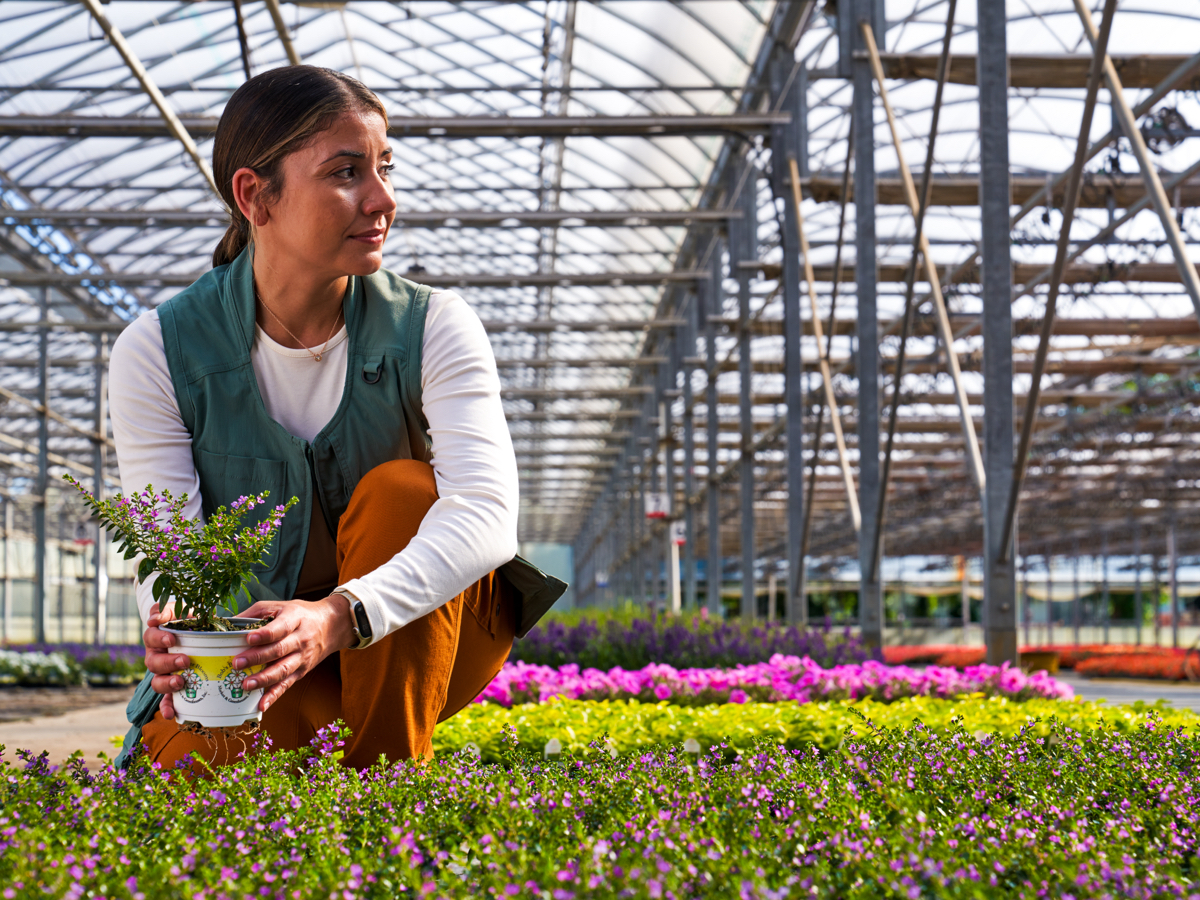 woman holding plant in greenhouse wearing green vest looking off to the side
